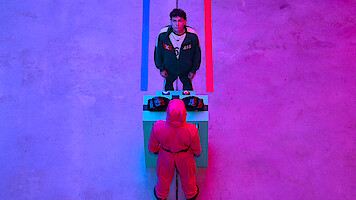 A man in a green tracksuit faces a masked figure in a pink jumpsuit across a table, under dramatic blue and pink lighting, creating a tense and surreal atmosphere in a minimalist setting.