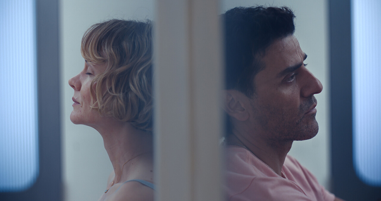 A woman and a man sit back-to-back separated by a wall in a modern, softly lit room, both appearing contemplative or distant, with subtle blue lighting in the background.