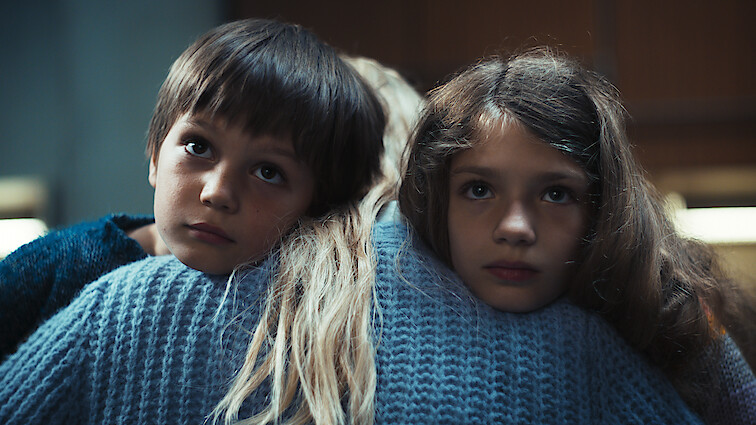 Two children, Jonathan and Hannah, face the camera, embraced by a woman whose faces away from the camera, her long blonde hair falling down her back. A still from 'Dear Child.'