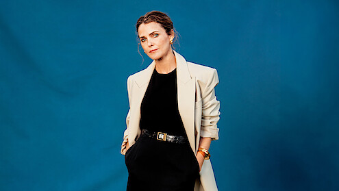 Woman posing against a solid blue background, wearing a light-colored blazer over a black outfit with hands in her pockets, accessorized with a belt and bracelet, looking relaxed and confident in a studio setting.