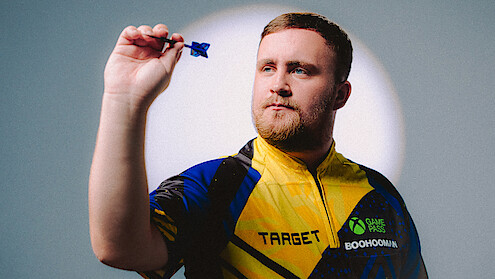 A man wearing a colorful dart shirt holds a dart up in a throwing position, focused, with a light spotlight background in a studio setting.