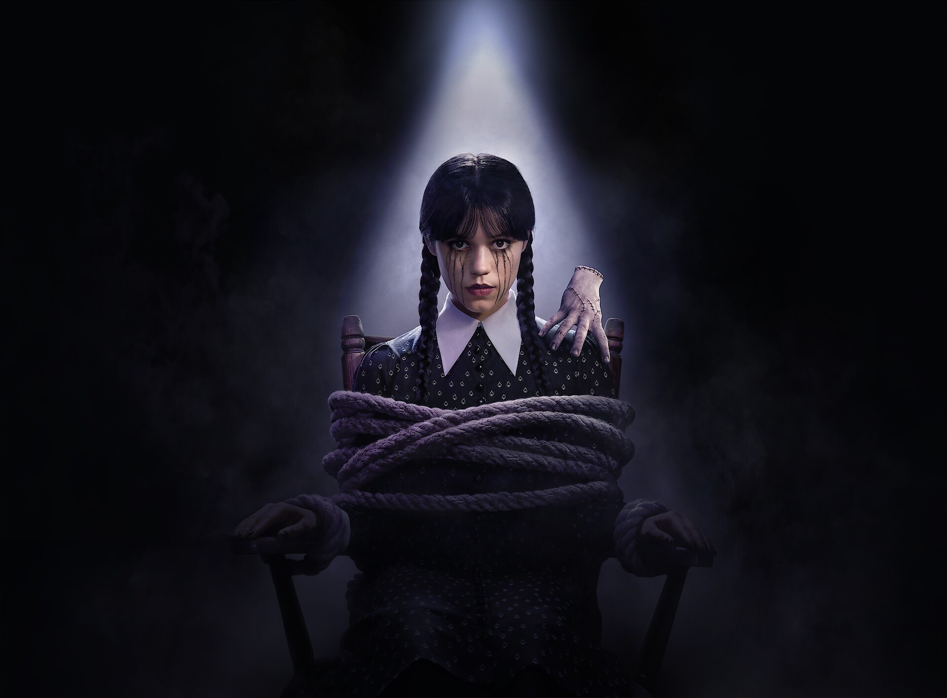Jenna Ortega as Wednesday, who is tied up in a chair with Thing on her shoulder. 