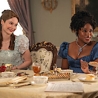 Two women in elegant period dresses sit at a lavishly set dining table in a richly decorated room, enjoying tea and desserts, with ornate chairs and a soft natural light coming through curtained windows.