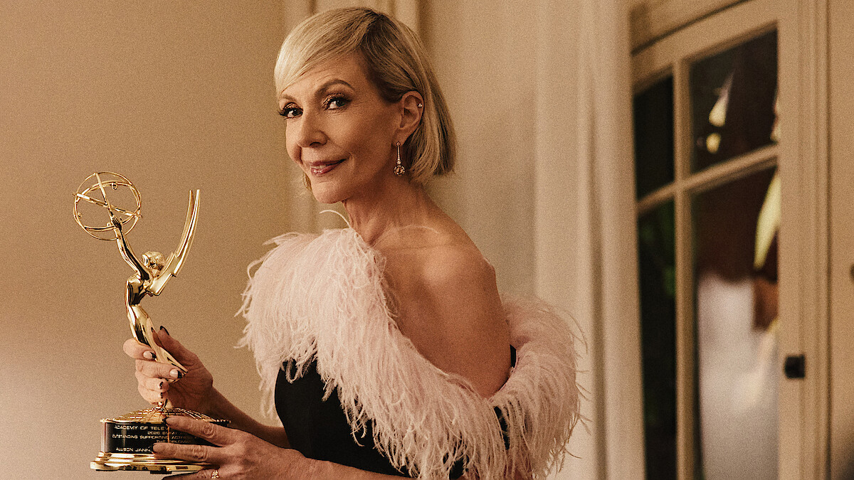 Woman in an elegant black dress with a pink feathered collar holding an Emmy award, standing indoors with warm lighting and a window in the background.