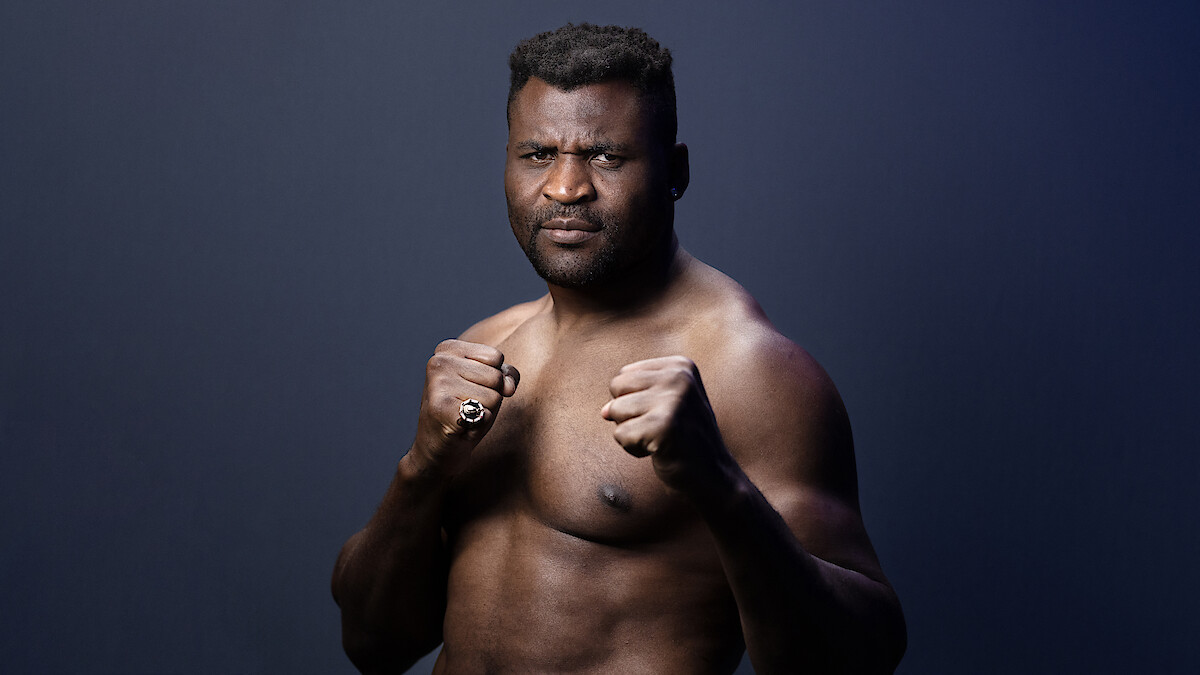 Francis Zavier Ngannou in a fighting stance with clenched fists, looking serious, posing against a plain dark gradient background in a studio setting.