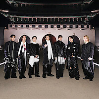 V, JIN, SUGA, RM, JUNG KOOK, J-HOPE, and JIMIN dressed in fashionable black and white outfits stand outdoors at night in front of an ornate, traditional East Asian building with arches and a large decorative roof.