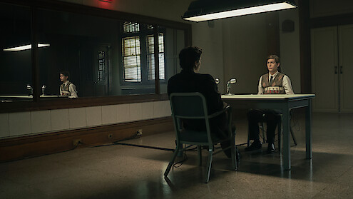 Two people sit across from each other at a table in a dimly lit interrogation room, with a large mirror on the wall reflecting the scene and a microphone on the table, creating a serious and tense atmosphere.