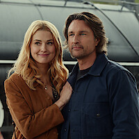 A smiling man and woman walk together outdoors, dressed in casual jackets, with a lush green background suggesting a park or natural setting.