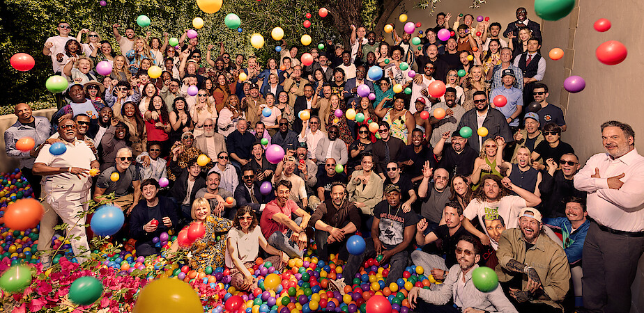 Large group of people of various ages gathered outdoors, posing in a ball pit with colorful plastic balls bouncing in the air; casual and joyful setting with trees and a wall in the background.