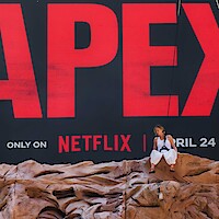 A person sits on artificial rocks in front of a large billboard for a Netflix show or movie, featuring bold red letters and a release date of April 24, creating a dramatic promotional scene.