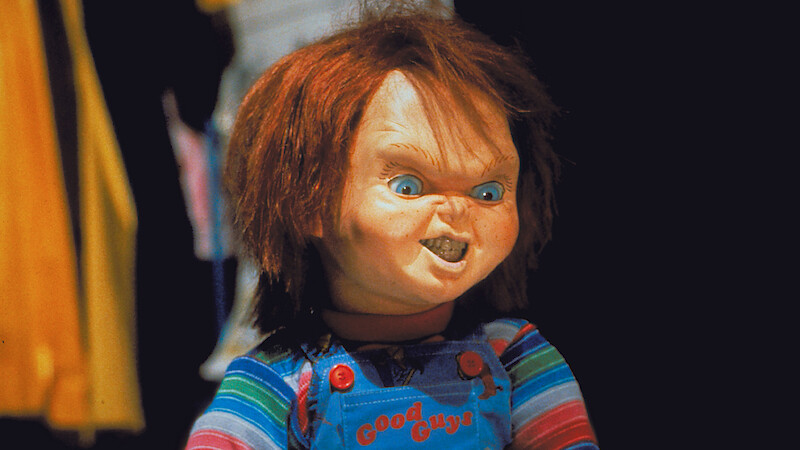 A creepy doll with red hair and overalls stands indoors, wearing a striped shirt. The background is dark with indistinct, colorful objects and clothing visible. The doll has a menacing facial expression.