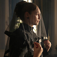 Woman in black mourning attire with a sheer veil over her face, standing indoors in soft light, holding the veil with one hand, looking thoughtful in a formal, vintage setting.