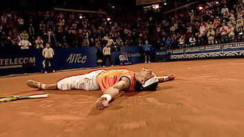 Tennis player lying on back on clay court, arms spread, appearing exhausted or celebrating victory, with tennis racket beside and cheering crowd in evening stadium.