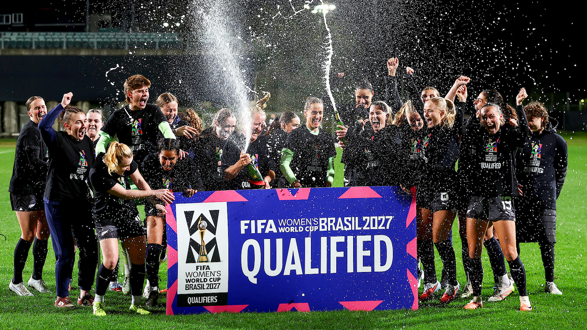 Women's soccer team celebrates at night on a field, spraying champagne and cheering behind a large sign reading "FIFA Women's World Cup Brasil 2027 Qualified." Bright stadium lights and festive atmosphere.