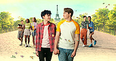 Joe Locke as Charlie and Kit Connor as Nick hold hands and walk down the streets of Paris with the cast of Season 2 of 'Heartstopper' behind them.