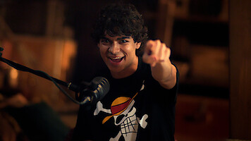 Iñaki Godoy smiles and points at camera, wearing a black t-shirt with a the Straw Hats Jolly Roger logo, sitting indoors in a warmly lit room with a microphone in front of him.
