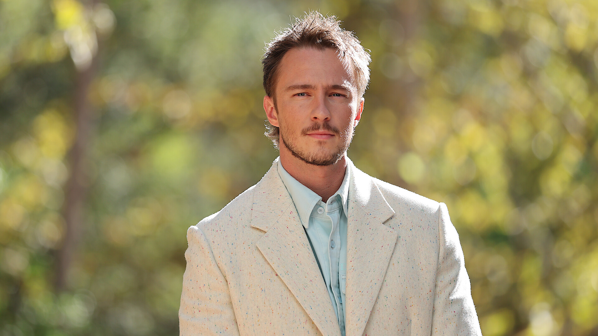 Drew Starkey in a light-colored suit stands outdoors with a blurred background of greenery and sunlight.