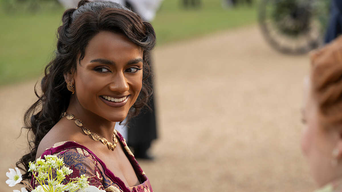 A woman in a period costume smiles outdoors, holding a bouquet of flowers, with people and a carriage blurred in the background, suggesting a historical or formal event setting.