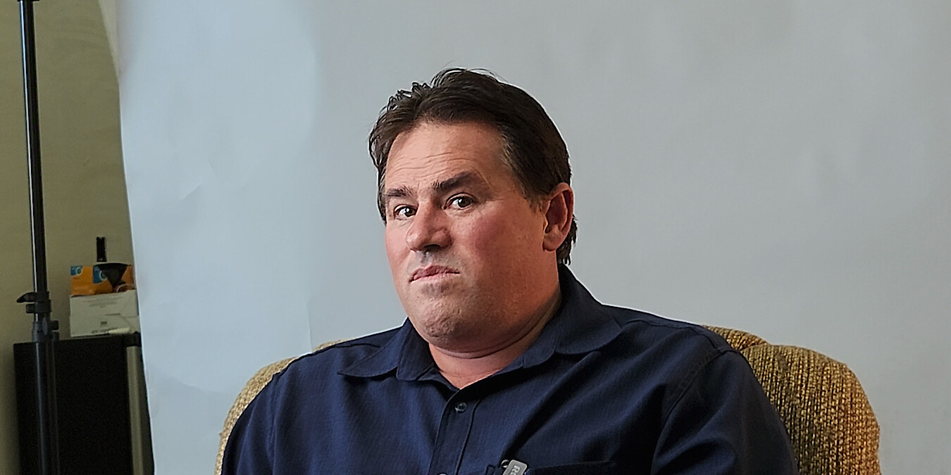 A man with brown hair in a blue collared shirt sits in front of a white background and looks directly to camera.