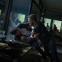 Two men struggle violently in an office, one pinning the other against a desk, while others watch in shock. The setting is a cluttered workspace with glass walls and scattered files, lit by daylight through window blinds.