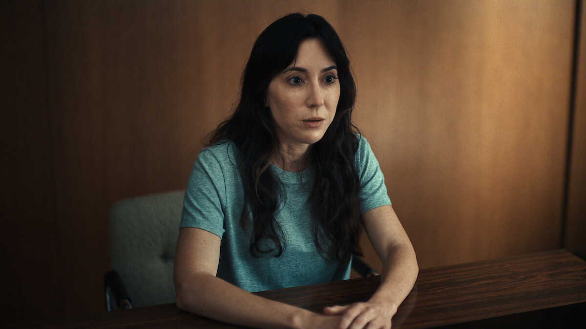Woman with long dark hair in a light blue t-shirt sitting at a wooden desk in a dimly lit, wood-paneled room, looking forward with a serious expression.