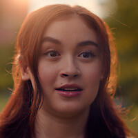 A young woman with long brown hair, outdoors in a natural, sunlit setting, looking surprised or intrigued. Blurred green background and warm lighting.