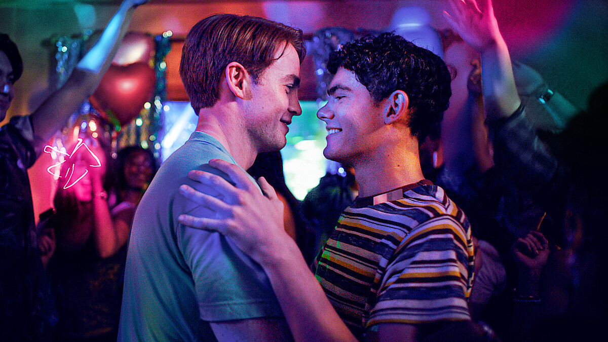 Two young men smile and embrace while dancing closely at a lively, colorful party, with people cheering and dancing in the vibrant, festive background.