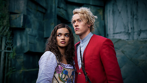 Two young adults in period costumes, a woman in a white dress with floral accents and a man in a red jacket, stand close together looking ahead, with a stone wall or ruins as the background, in a dramatic, moody setting.