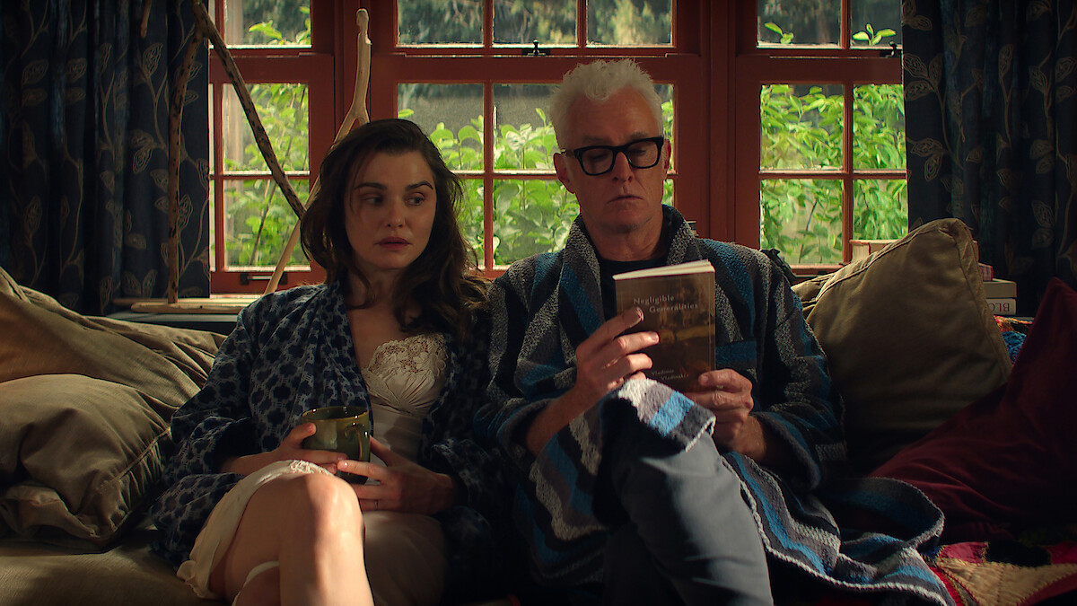 Rachel Weisz as M and John Slattery as John in bathrobes sit on a cozy couch by large windows, one holding a drink, the other reading a book, surrounded by cushions and natural light, with greenery visible outside.