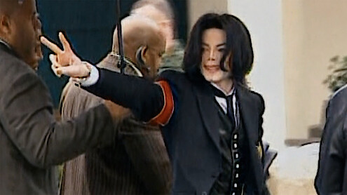 Person in a black suit with red armband making a peace sign outdoors, surrounded by several people in a public or official setting.
