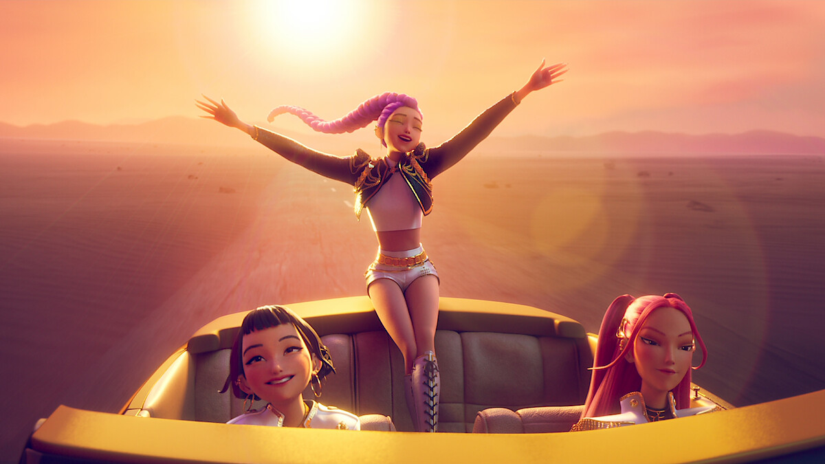 Three animated girls in futuristic outfits ride in a convertible through a desert at sunset; one stands with arms outstretched, while the others smile and sit in the car, golden sunlight in the background.