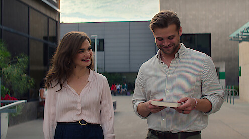 Rachel Weisz and Leo Woodall walk across campus. Woodall reads the back of a book, as both smile.