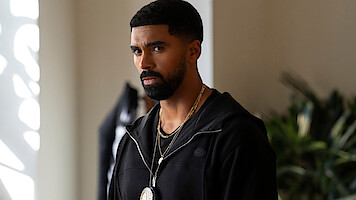A serious man with a trimmed beard and short hair, wearing a black jacket and a badge on a chain around his neck, stands indoors near plants with light streaming in from a window.