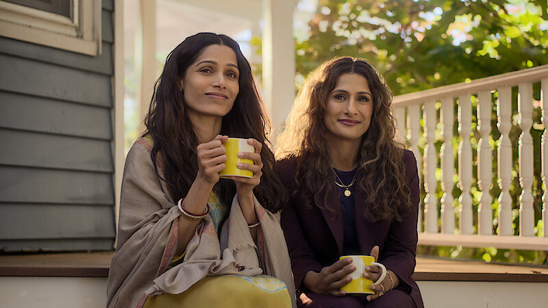 Two women sit together on a sunlit porch, holding yellow mugs and smiling. Shrubs and a white railing are visible in the background, creating a relaxed, inviting environment.