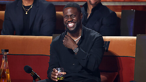 Man in black shirt sitting on a couch at an event, laughing, holding a drink, with a microphone and bottles on the table in front of him, and other people seated in the background.