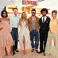 Six people dressed in formal and semi-formal attire stand together smiling on a tan carpet at a Netflix "Outer Banks" event, with a bright orange and yellow promo sign in the background outdoors.