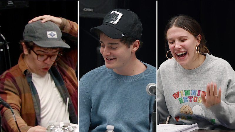Gaten Matarazzo, Noah Schnapp, and Millie Bobby Brown sit at a table with scripts and microphones, laughing and reacting animatedly, in a casual, creative studio environment suggesting a lively script reading or rehearsal session.