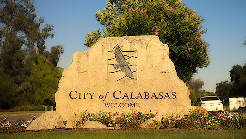 Large stone welcome sign for the City of Calabasas with an eagle engraving, surrounded by flowers, trees, and parked cars in a sunny outdoor setting.