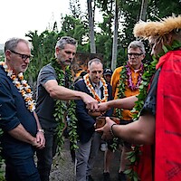 A group of men wearing leis and greenery participate in a traditional outdoor ceremony with a cultural leader, surrounded by tropical trees and greenery.