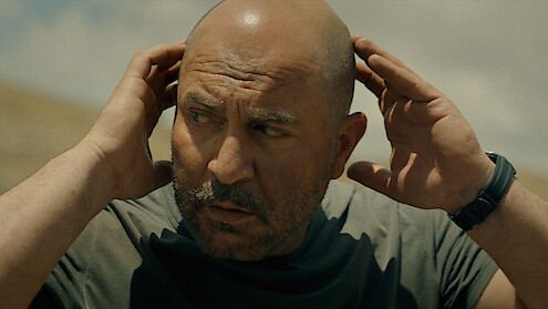 Bald man outdoors under cloudy sky, wearing a gray t-shirt and a watch, holding both hands to his head, looking concerned or focused in a natural setting.