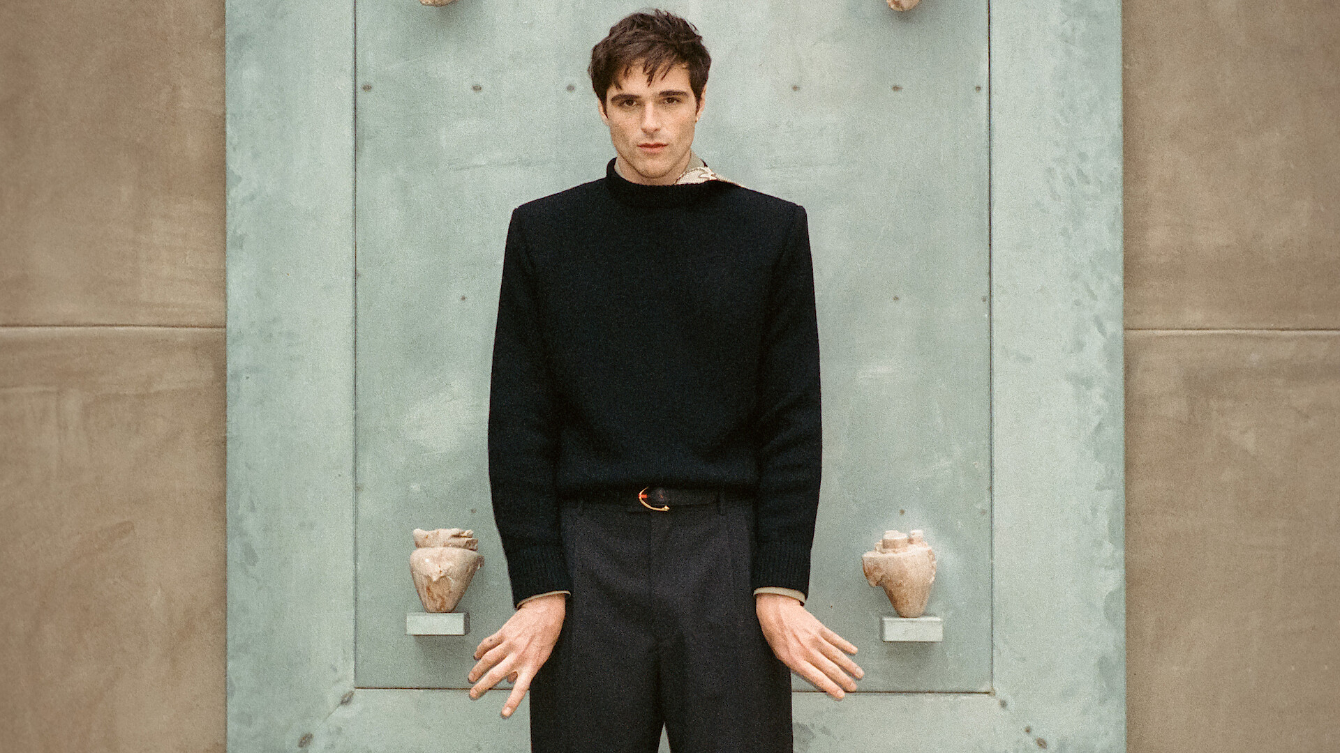 Jacob Elordi stands in front of an artwork featuring anatomically correct replicas of human hearts wearing a black sweater, trousers, belt, and shoes.