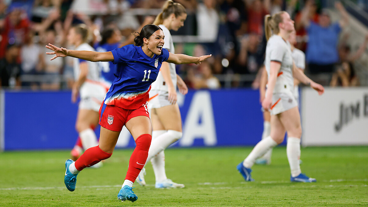 Soccer player in blue jersey celebrates with arms outstretched on a stadium field, surrounded by other players in white uniforms and a cheering crowd in the background during a match.