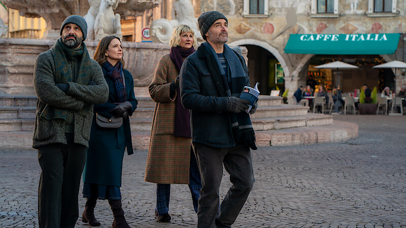 Four adults in winter clothing walk together in a European plaza near a fountain, with a café and people dining outside in the background, suggesting a casual sightseeing outing in an urban historic setting.