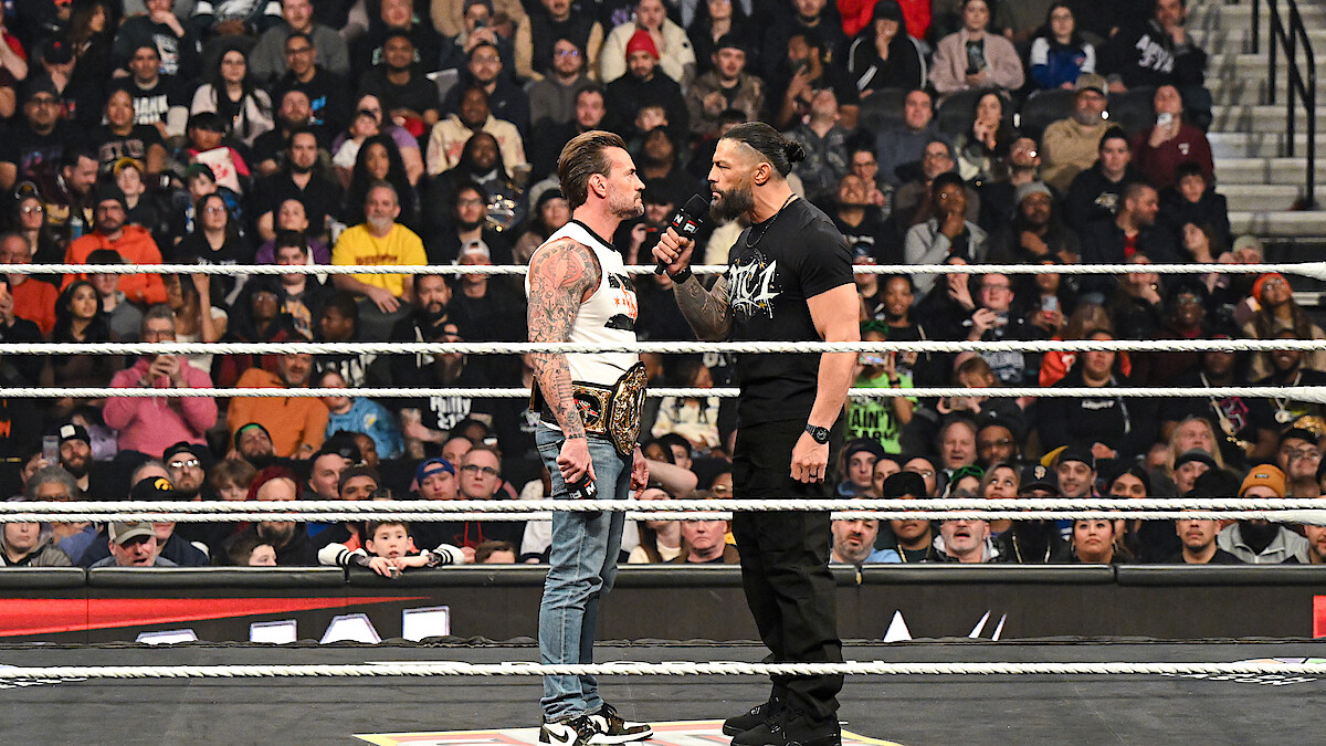 Two wrestlers face off in a packed arena wrestling ring, one holding a championship belt and the other speaking into a microphone as a large audience watches the dramatic confrontation.