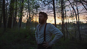 A man stands in a forest at dusk with hands on hips, surrounded by tall trees and fading sunlight, creating a calm and reflective mood.