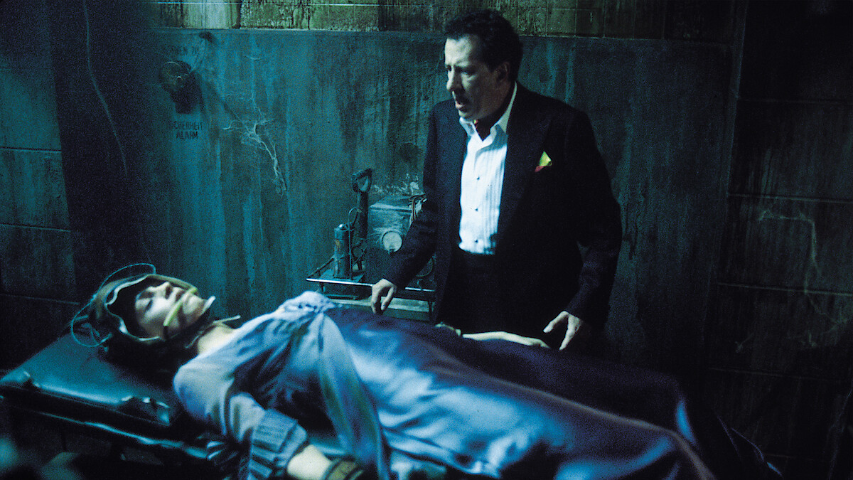 A man in a suit stands in a dimly lit, rundown room next to a restrained person lying on a table, creating a tense and dramatic atmosphere suggestive of a suspense or horror setting.