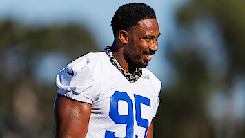 Football player wearing a white jersey with the number 95 smiles during an outdoor practice session, with blurred trees and blue sky in the background.