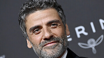 Man with curly hair and beard in formal attire smiles at a black tie event, standing in front of a dark backdrop with white decorative text and floral graphics.