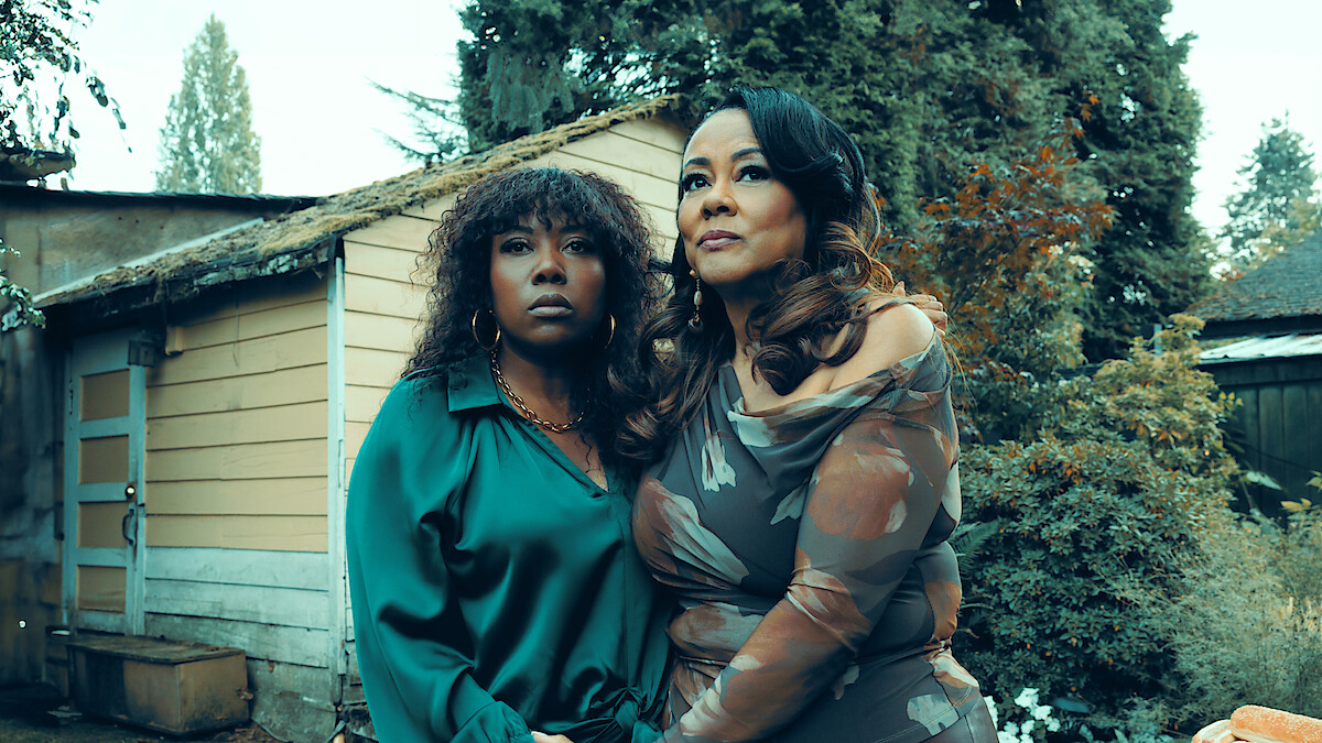 Two women stand close together outside beside a rustic shed, surrounded by greenery and trees. Both look thoughtful, dressed formally, with overcast natural lighting creating a serene, introspective mood.