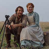 A man and woman in pioneer clothing sit together outdoors on grass, next to a campfire tripod and a basket, with dry prairie landscape in the background under a clear sky.
